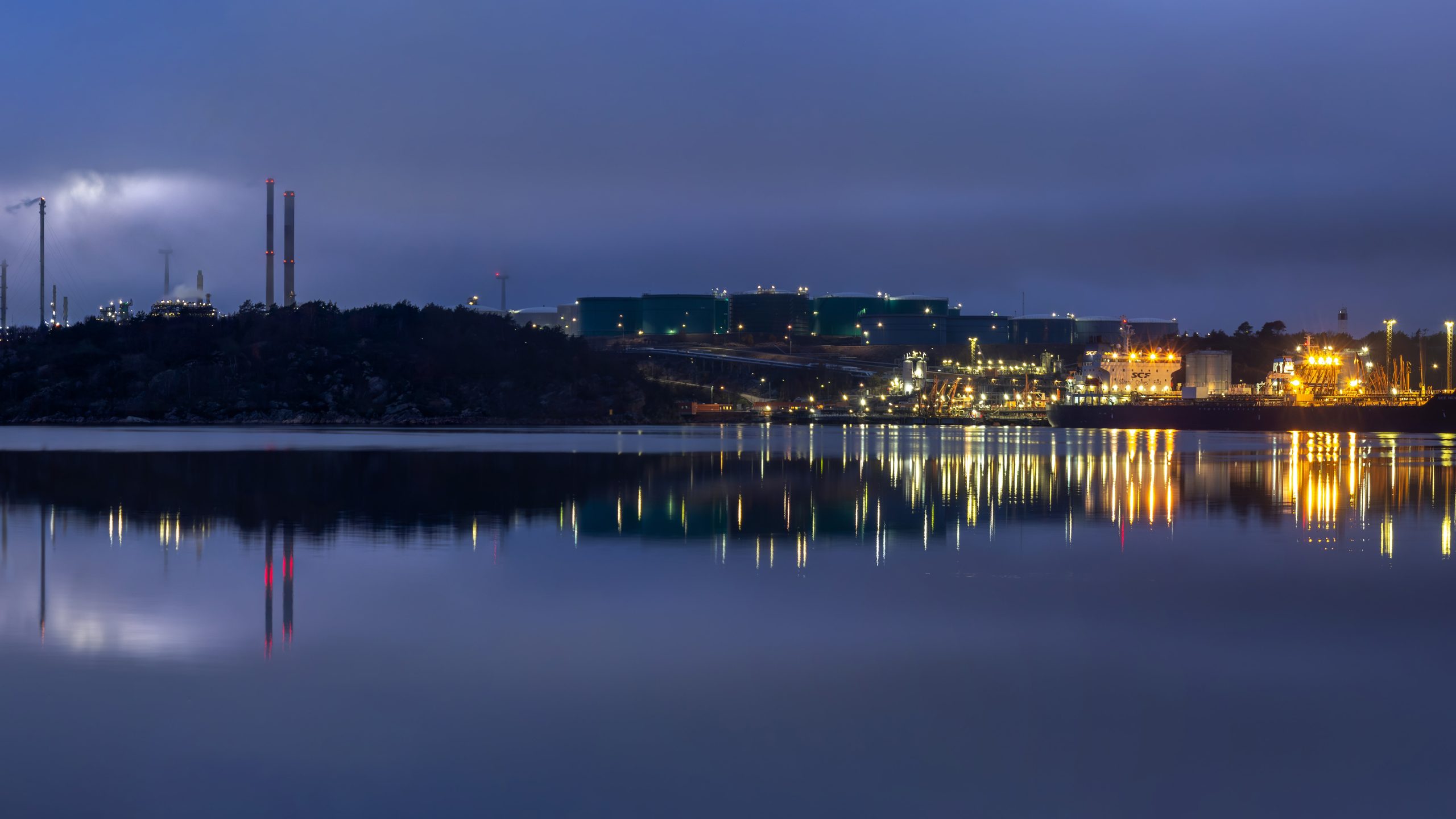 Silhouette of an oil refinery with smokestacks against a blue-hour sky.