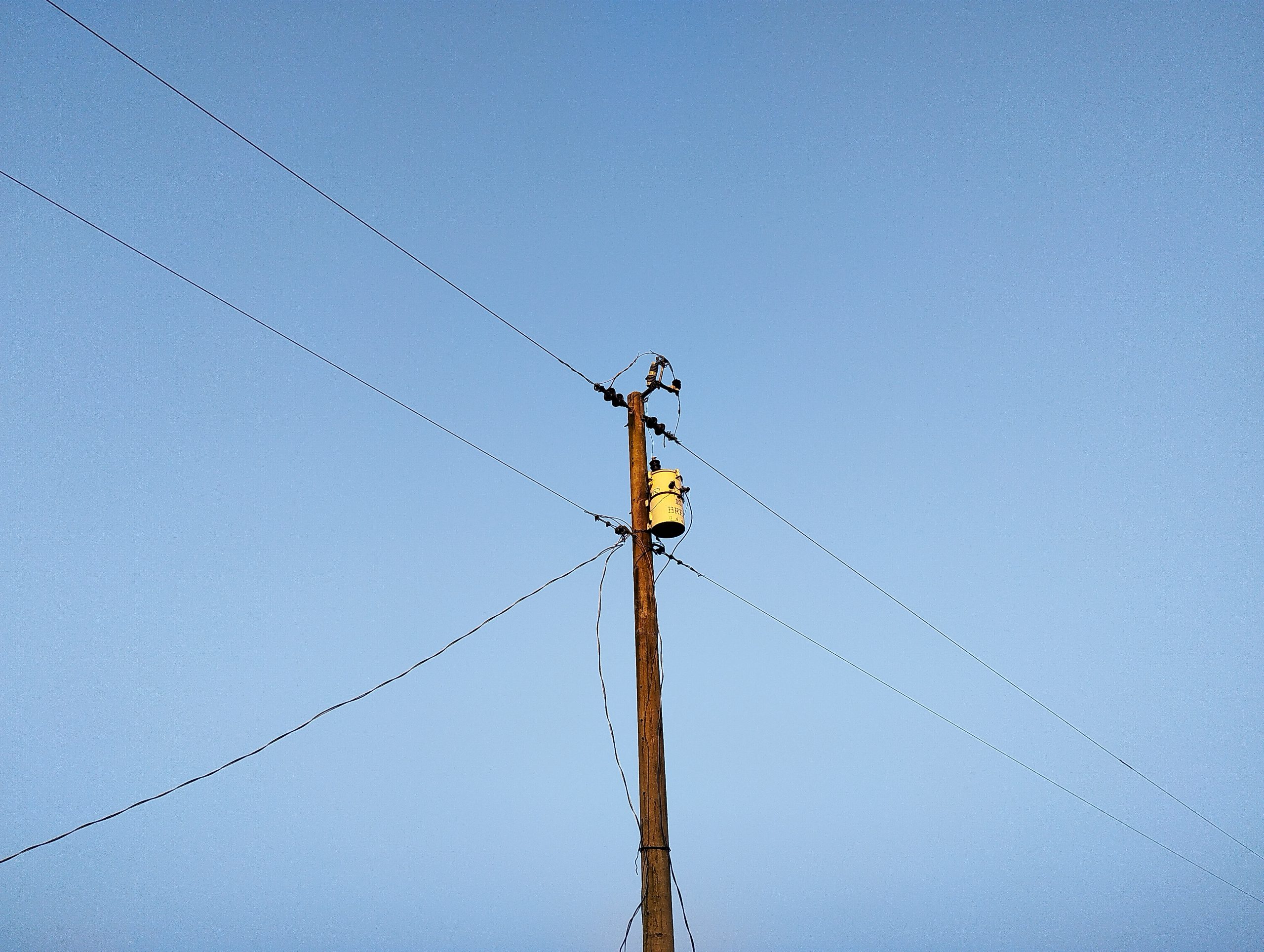 A rural Bangladesh wooden power-line pole with transformer against a blue sky.